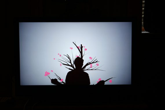 A person in silhouette form standing in front of an interactive and illuminated screen which features branches and pink flowers blooming from their silhouette.