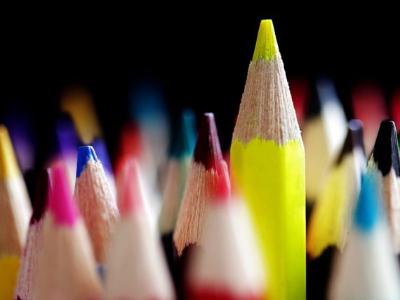 A close up image of coloured pencils, with one pencil in yellow, slightly elevated above the rest. Set on a black background.