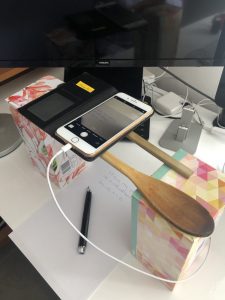 An iPhone held above a piece of paper by two wooden kitchen utensils propped on top of tissue boxes.