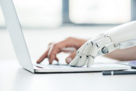 one human and one robot hand typing on a laptop