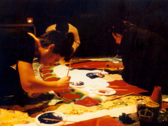 a moody shot with black and red tones and low, warm lighting, shows three people sitting on a large piece of cloth on which they’re painting an artwork together