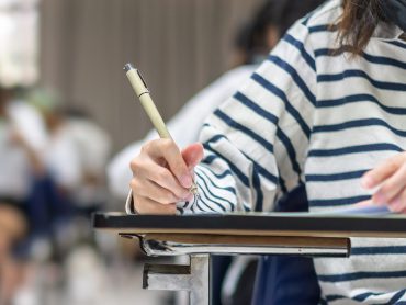 An exam hall with students sat at desks completing an exam with pen and paper. A female student in a blue and white striped sweater, face out of view, is in the foreground and holding a beige pen. Students are seen in the background.