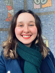 Profile image of a woman wearing a teal green coat and forest green scarf. She has shoulder length light brown hair, blue eyes, and is smiling at the camera. She is standing against a blue mosaic tiled background. 