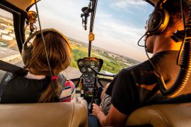 A child with long hair tied back in a pony tail and a man with short hair fly a helicopter together, we look out the front of the helicopter and can see the controls and the view and the back of their heads