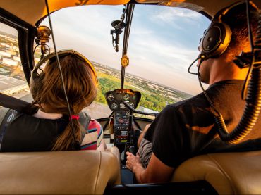 A child with long hair tied back in a pony tail and a man with short hair fly a helicopter together, we look out the front of the helicopter and can see the controls and the view and the back of their heads