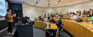 A lecture theatre filled with people is receiving a presentation from a young man with black hair and a tan jacket. 