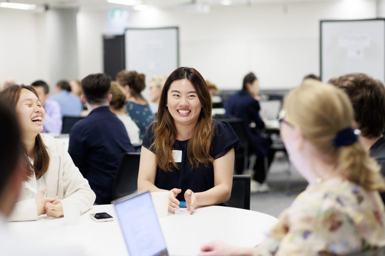 Students and staff sit around a tables in a conference room. At the centre of the image a student with long dark hair and a black t-shirt smiles in conversation with a woman with blonde hair sat on the other side of the table.