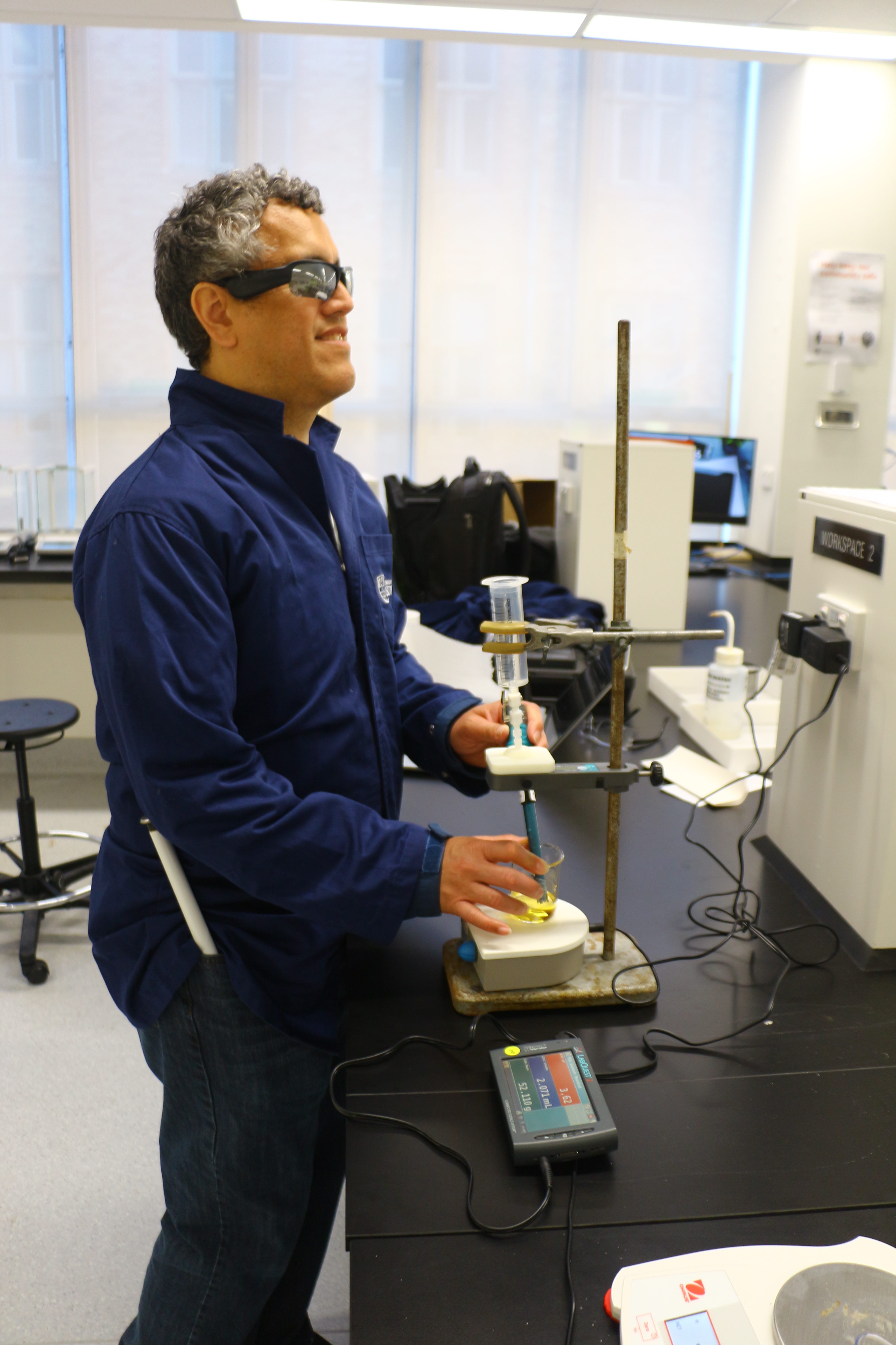 IMAGE DESCRIPTION: Image is a photograph of Dr Cary Supalo, which shows him wearing safety glasses and trialling a titration experiment in the 1st Year Chemistry Laboratory at USyd. Cary is holding a beaker containing a yellow solution to which a substance is being added dropwise. A pH sensor in the beaker is attached to a Talking LabQuest2 that reads out the pH reading.