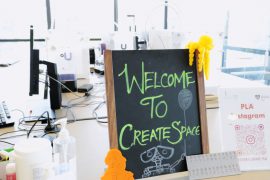 Close up of little blackboard with green chalk saying 'Welcome to create space', with a 3d printed yellow octopus and orange Shakespeare near it, on a table with computers, and happy clutter