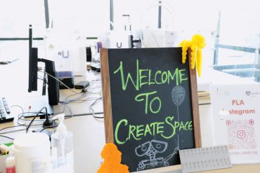 Close up of little blackboard with green chalk saying 'Welcome to create space', with a 3d printed yellow octopus and orange Shakespeare near it, on a table with computers, and happy clutter