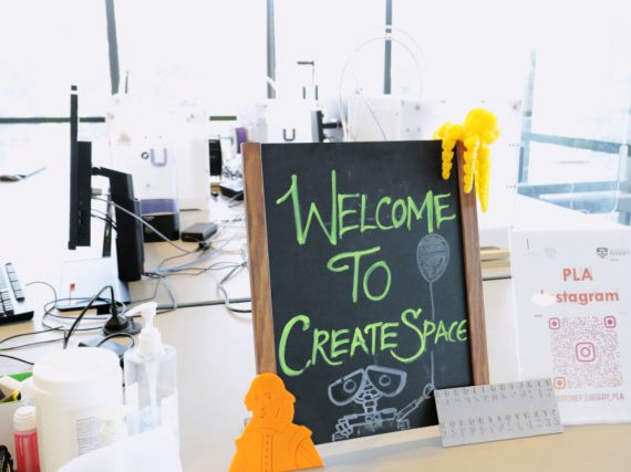 Close up of little blackboard with green chalk saying 'Welcome to create space', with a 3d printed yellow octopus and orange Shakespeare near it, on a table with computers, and happy clutter