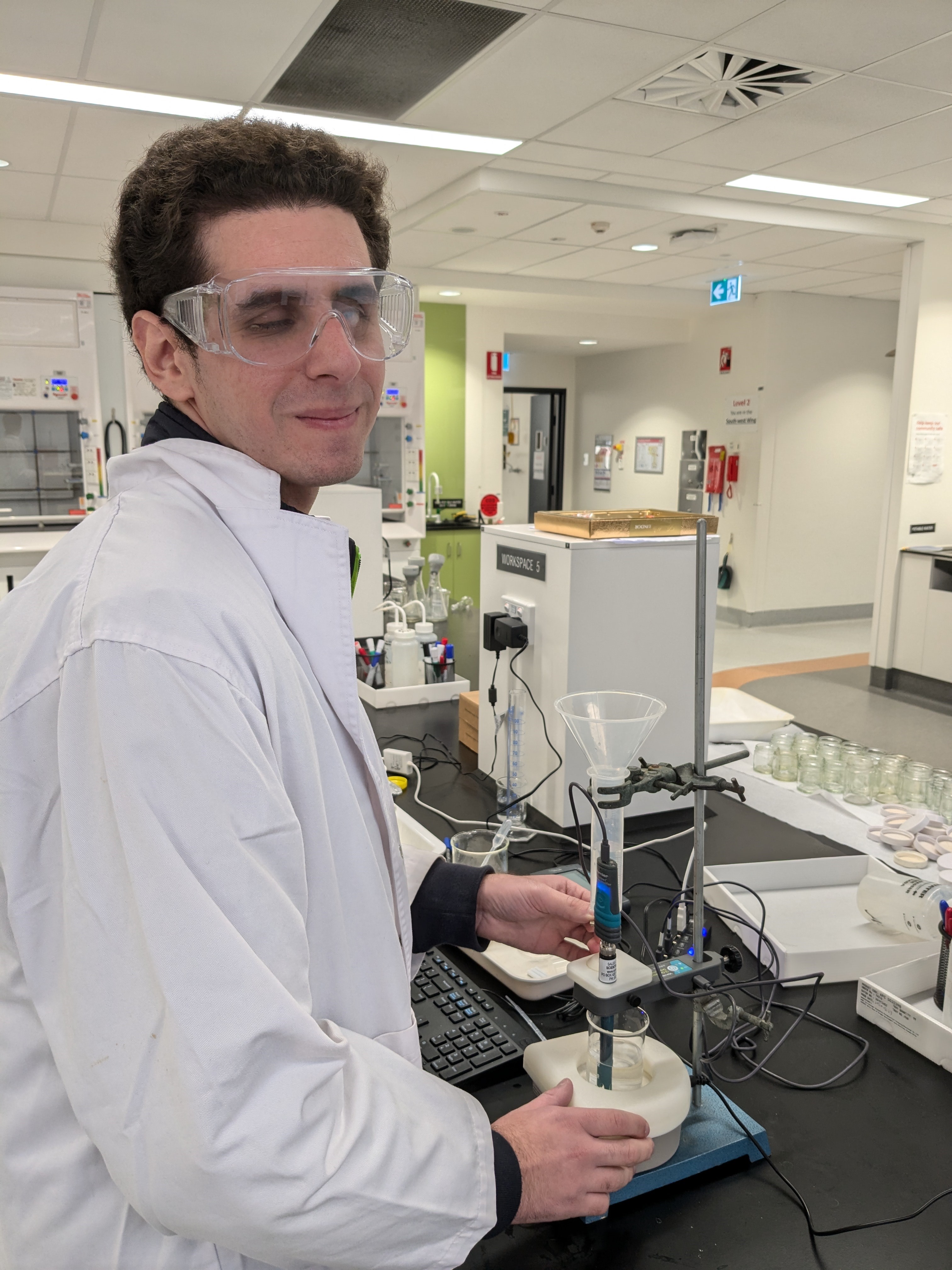 IMAGE DESCRIPTION: Image is a photograph of Mr Theodore Tsolakis in the first year chemistry labs at USyd . Theodore wears a labcoat and safety classes and is using a Talking LabQuest 2 to trial the experimental procedures developed at USyd.