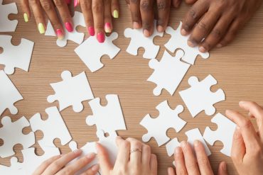 A panoramic overhead view of multiple hands from diverse individuals working together to assemble a jigsaw puzzle against a white background. The hands are reaching from different directions to connect puzzle pieces, demonstrating teamwork and collaboration.