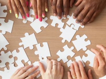 A panoramic overhead view of multiple hands from diverse individuals working together to assemble a jigsaw puzzle against a white background. The hands are reaching from different directions to connect puzzle pieces, demonstrating teamwork and collaboration.