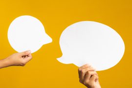 Two hands holding empty white speech bubbles against a yellow background. Close-up photo.