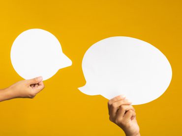 Two hands holding empty white speech bubbles against a yellow background. Close-up photo.