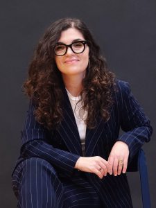 Profile image of a Dr Laura Crocco, sat down wearing a dark blue pin-striped suit with a white t-shirt. She is smiling at the camera and has long dark hair worn down and a pair of black framed glasses.