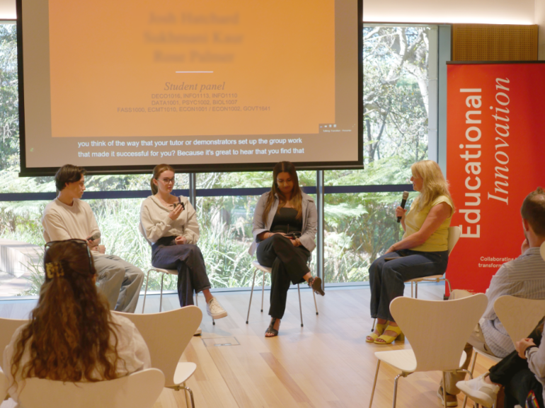 Student panel and MC sit speaking at the End of Year event in front of a slide screen with a big window and garden beyond.