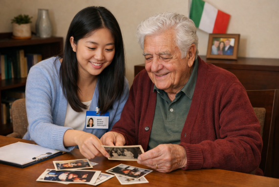 A speech pathology student of Asian background sits beside an elderly man of Italian background at a table, smiling as they look through printed photographs together. The student gently points to one photo while the man holds another, both appearing engaged and happy. A clipboard and more photos rest on the table, suggesting a reminiscence activity in a warm, home-like setting.